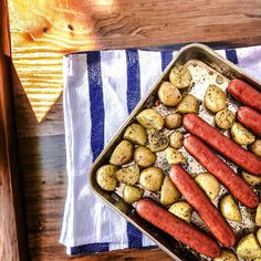 a pan filled with hot dogs and potatoes on top of a towel next to a wooden table