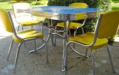 a blue table and four yellow chairs sitting on the sidewalk in front of some bushes