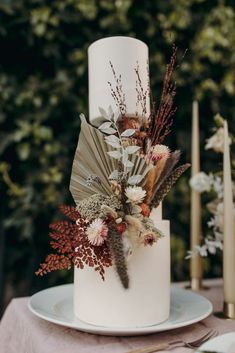 a white wedding cake with dried flowers and feathers on the top, surrounded by candles