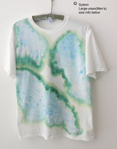 a white t - shirt with green and blue paint on it hanging from a hanger