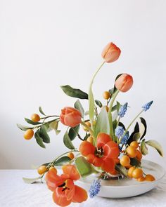 an arrangement of flowers in a bowl on a white table cloth with leaves and berries