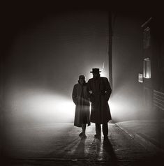 two men standing in the middle of a street at night, one wearing a long coat and hat