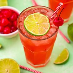 two glasses filled with red and green drinks next to limes, cranberries, and cherries