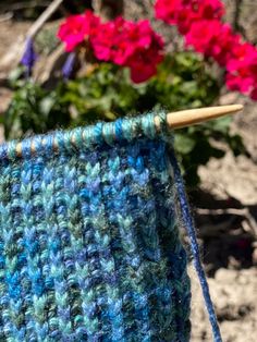 a blue and green knitted object next to some pink flowers in the background with a wooden stick sticking out of it