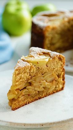 a slice of apple cake on a plate with another piece in the background and two green apples behind it