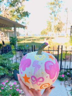 a hand holding up a painted pumpkin in front of a fence and flowerbeds