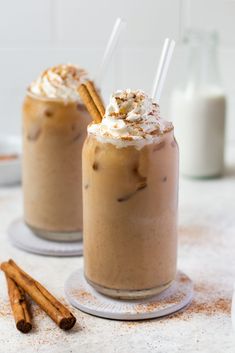 two glasses filled with hot chocolate drink and topped with whipped cream, cinnamon sticks and powdered sugar