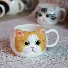 two mugs with cats painted on them sitting on a lace tablecloth next to each other