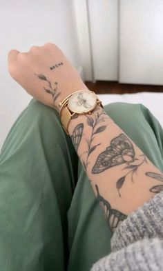 a woman's arm with tattoos and a watch on her left wrist, sitting in front of a white wall