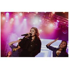 two women on stage singing into microphones with bright lights behind them and purple lighting