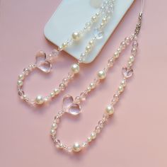 two necklaces with pearls are on a pink surface next to a cell phone and wallet