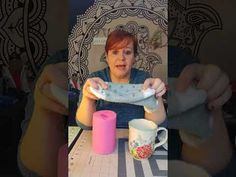 a woman sitting at a table holding up a piece of cloth next to two mugs