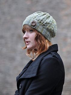 a woman wearing a knitted hat with buttons on the front and side, standing in front of a brick wall