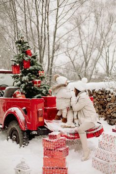 two people sitting on the back of a red truck with a christmas tree in the bed