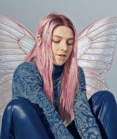 a woman with pink hair sitting on the ground wearing blue pants and a butterfly wings costume