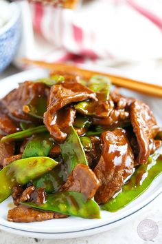 a white plate topped with stir fry beef and green peppers next to chopsticks