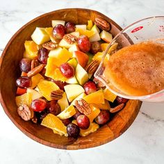 a wooden bowl filled with fruit and nuts next to a glass of orange juice on top of a marble counter