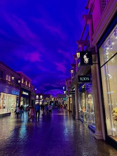 people are walking down the street in front of shops at night with purple clouds overhead