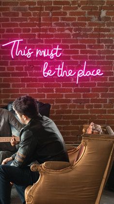 a man sitting on a couch in front of a brick wall with a neon sign