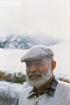 an old man with a white beard wearing a hat and plaid shirt standing in front of snow covered mountains