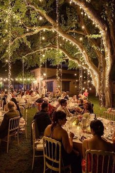 people are sitting at tables under trees with lights strung from the branches and around them