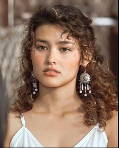 a woman with curly hair wearing earrings and a white top is looking at the camera