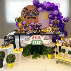 a party table with purple and yellow balloons on the wall, potted plants and decorations