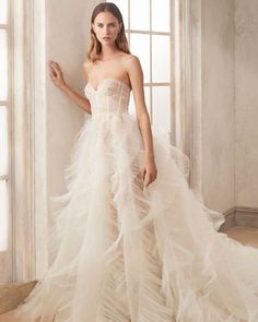 a woman in a strapless wedding dress with feathers on the skirt is standing by a window