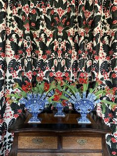 two blue vases with red flowers sit on a dresser in front of a floral wallpaper