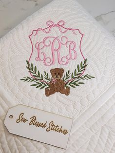 a teddy bear is sitting on top of a white quilted pillow with the initials monogrammed