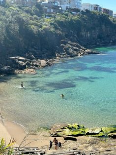 people are swimming in the clear blue water near some cliffs and houses on a hill