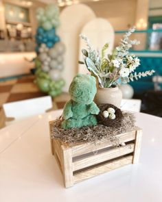 a small green teddy bear sitting on top of a wooden box next to a vase filled with flowers