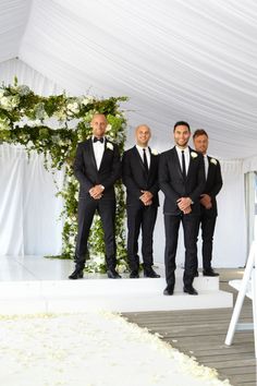 three men in tuxedos are standing under a white canopy at a wedding ceremony