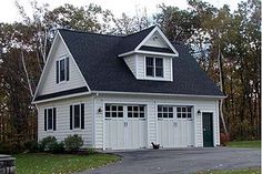 a white house with black shingles and two garage doors on the front, surrounded by trees