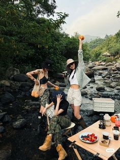 three women sitting at a picnic table on the side of a river, one holding an orange