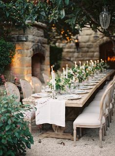 a long table with white linens and candles on it is set for an outdoor dinner