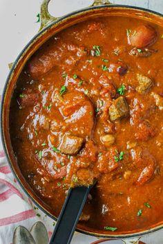 a pot filled with meat and vegetables on top of a white table cloth next to silver spoons