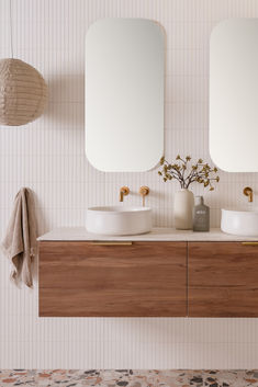 two white sinks sitting on top of a wooden cabinet next to mirrors and vases