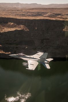 a fighter jet flying over a lake in the middle of mountains and desert land behind it