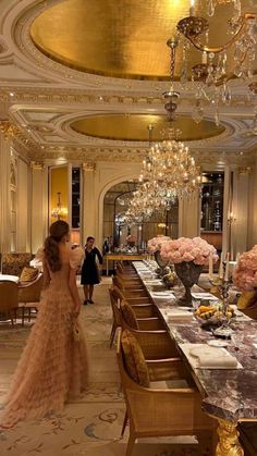 a woman standing in front of a long table with chairs and chandeliers hanging from the ceiling