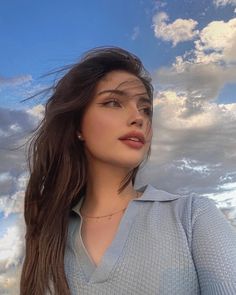 a woman with long hair standing in front of the sky wearing a sweater and earrings