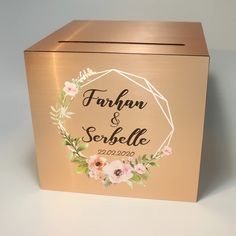 a gold box with floral wreath on the front and name written in black ink, sitting on a white surface