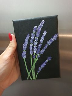 a woman's hand holding up a small square painting with lavender flowers on it