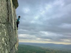 a man climbing up the side of a cliff on top of a mountain under a cloudy sky