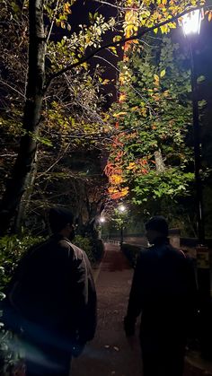 two people walking down a path in the woods at night with street lights shining on them