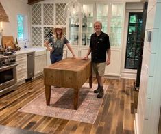 a man and woman standing in a kitchen next to a wooden table with a bench on it