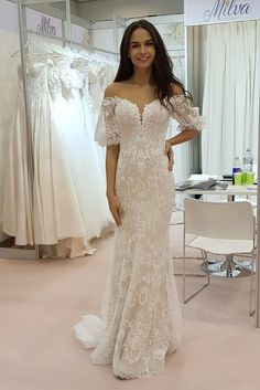 a woman standing in front of a dress on display at a bridal shop wearing an off the shoulder wedding gown