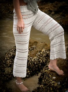 a woman in white pants standing on rocks by the water with her legs crossed and wearing sandals