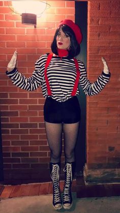 a woman dressed in black and white striped shirt with suspenders and red hat standing next to brick wall