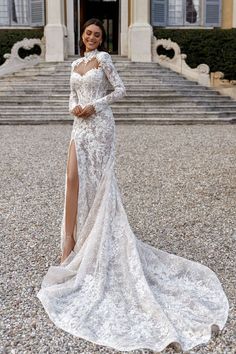 a woman standing in front of a building wearing a long sleeved wedding dress with high slit